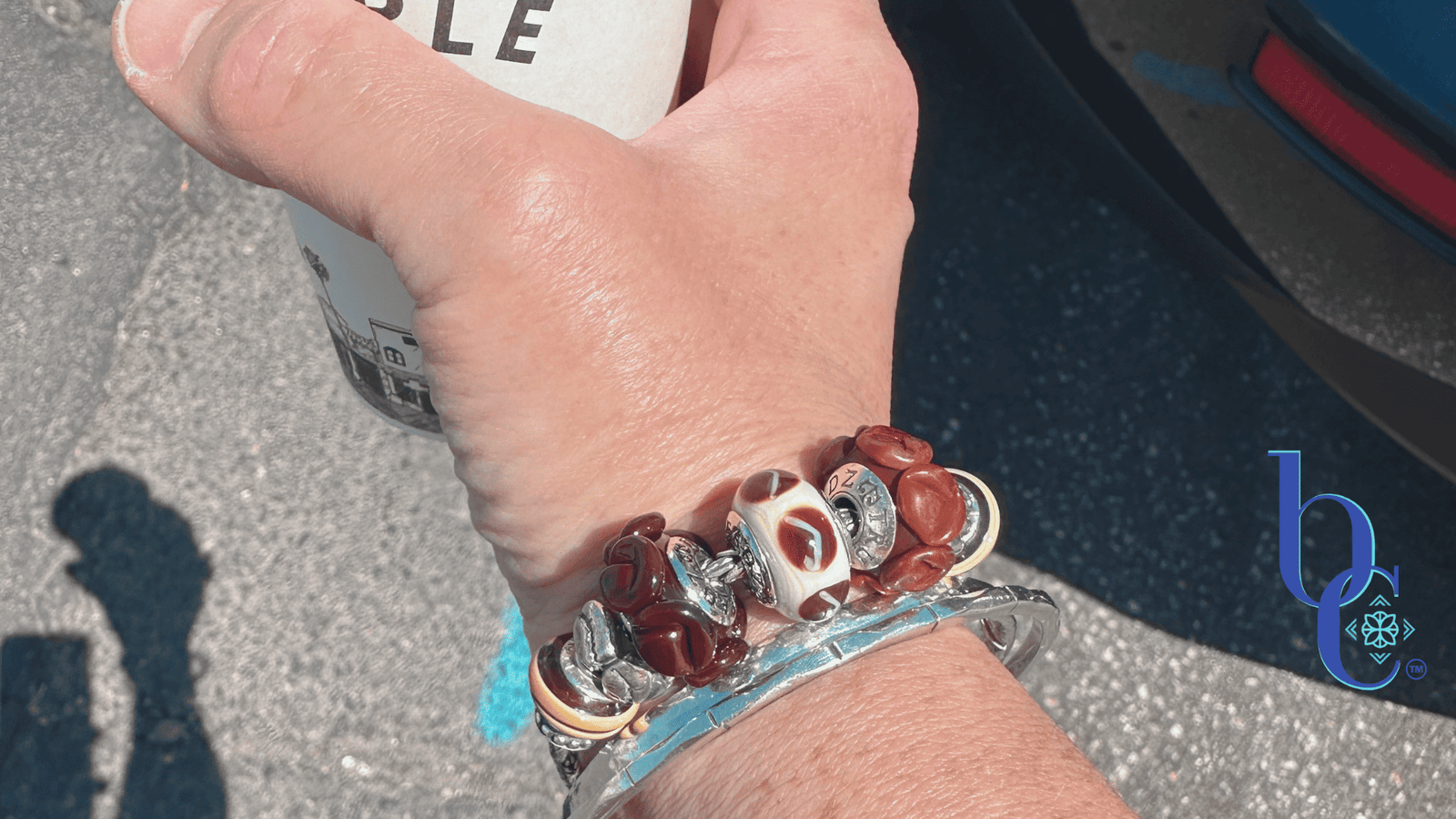 A woman's wrist wearing a chunky coffee-themed charm bracelet with TrueBeadz and Biagi Italy glass over a raw asphalt background.