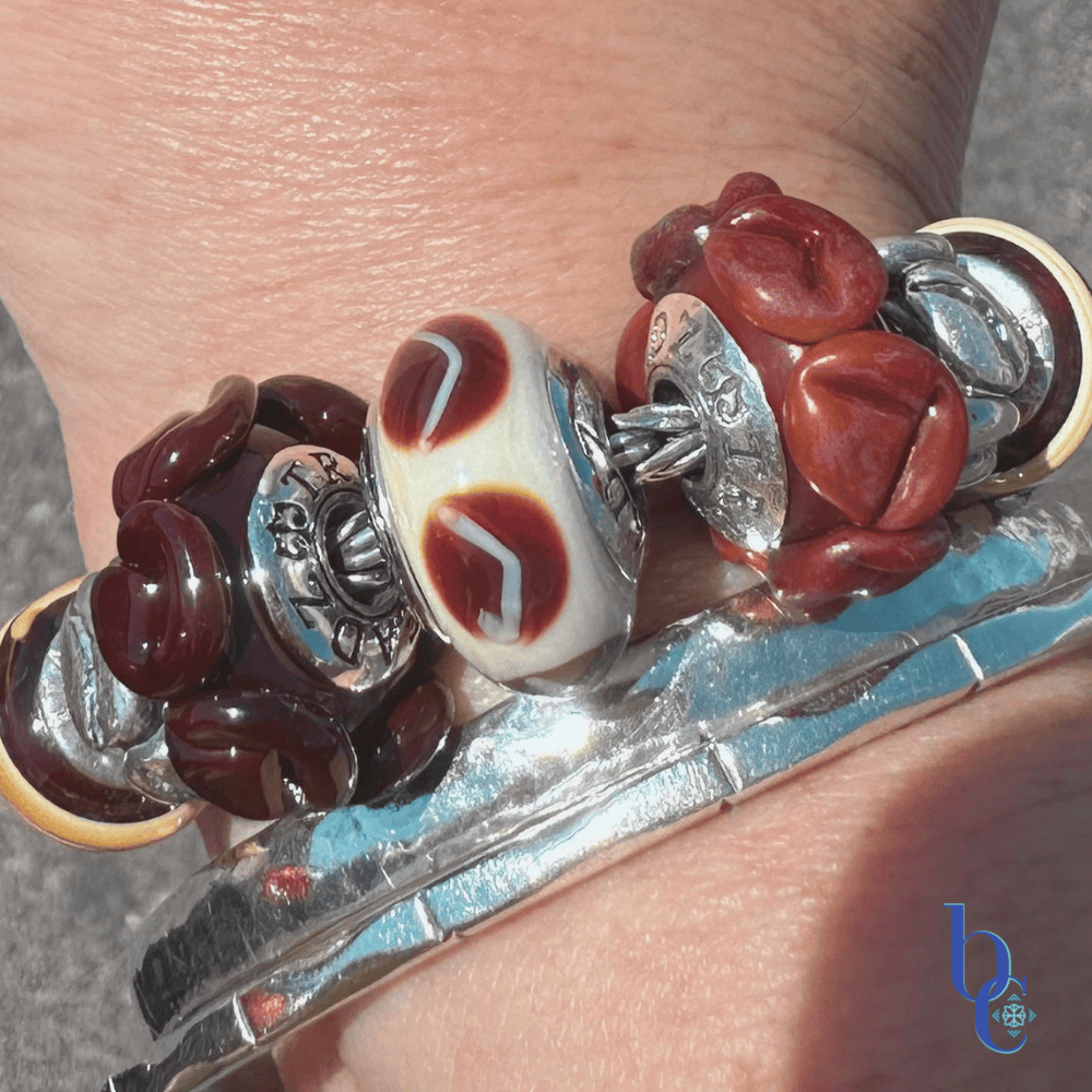 A woman's wrist wearing a chunky coffee-themed charm bracelet with TrueBeadz and Biagi Italy glass over a raw asphalt background.