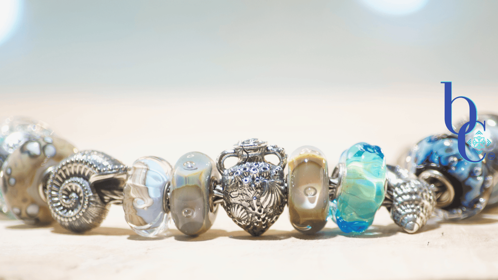 Close-up macro photograph of a European-style charm bracelet resting on a light surface. The focal beads include a textured silver nautilus shell, an aqua blue glass bead with internal bubbles, and the Kamane "Sandprint" bead featuring dark paw prints over a swirl of sandy beige and blue glass. The background is a soft, out-of-focus watercolor blue with bokeh lighting.