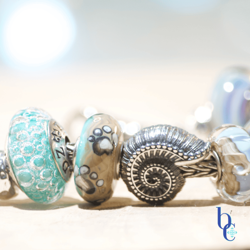 Close-up macro photograph of a European-style charm bracelet resting on a light surface. The focal beads include a textured silver nautilus shell, an aqua blue glass bead with internal bubbles, and the Kamane "Sandprint" bead featuring dark paw prints over a swirl of sandy beige and blue glass. The background is a soft, out-of-focus watercolor blue with bokeh lighting.
