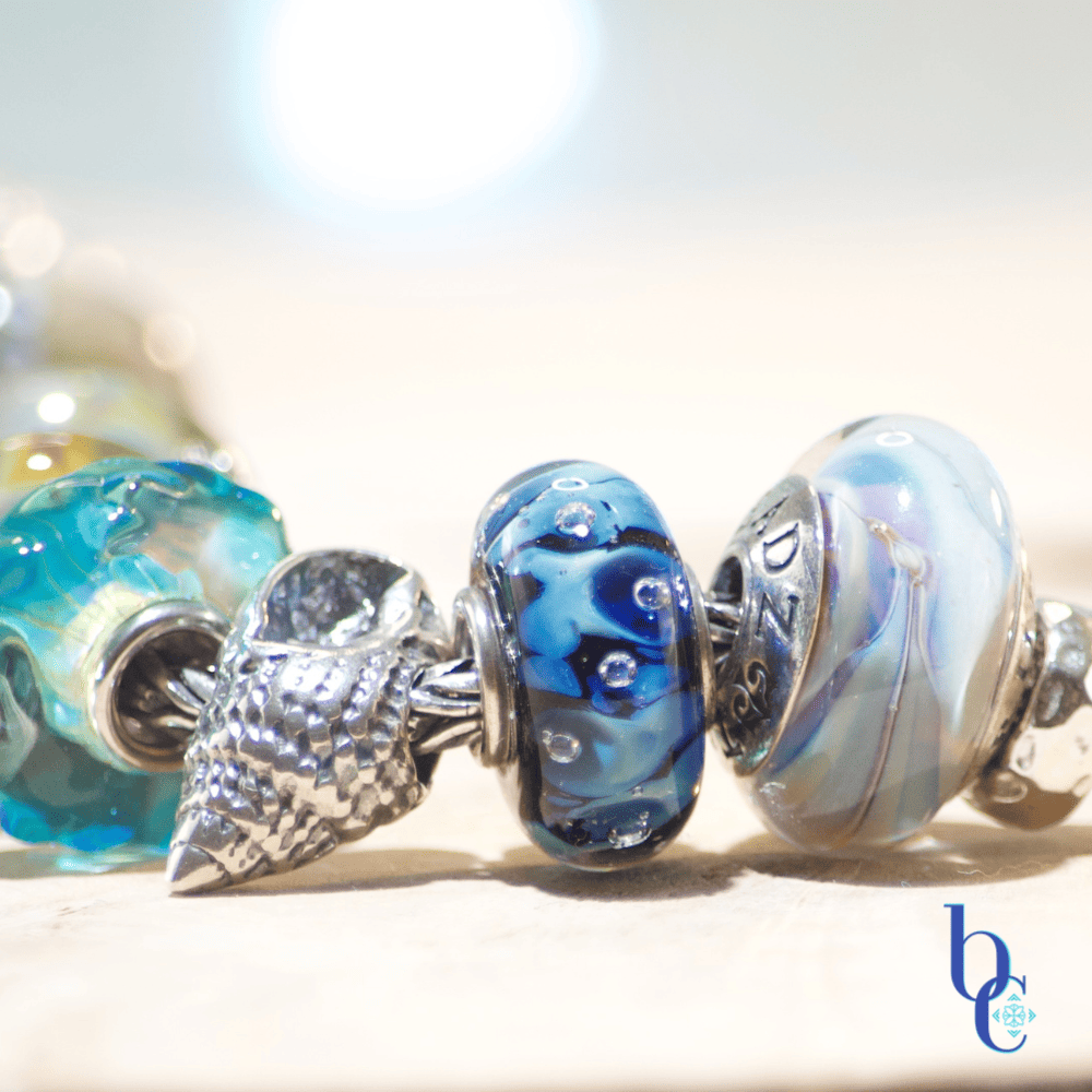 Close-up macro photograph of a European-style charm bracelet resting on a light surface. The focal beads include a textured silver nautilus shell, an aqua blue glass bead with internal bubbles, and the Kamane "Sandprint" bead featuring dark paw prints over a swirl of sandy beige and blue glass. The background is a soft, out-of-focus watercolor blue with bokeh lighting.
