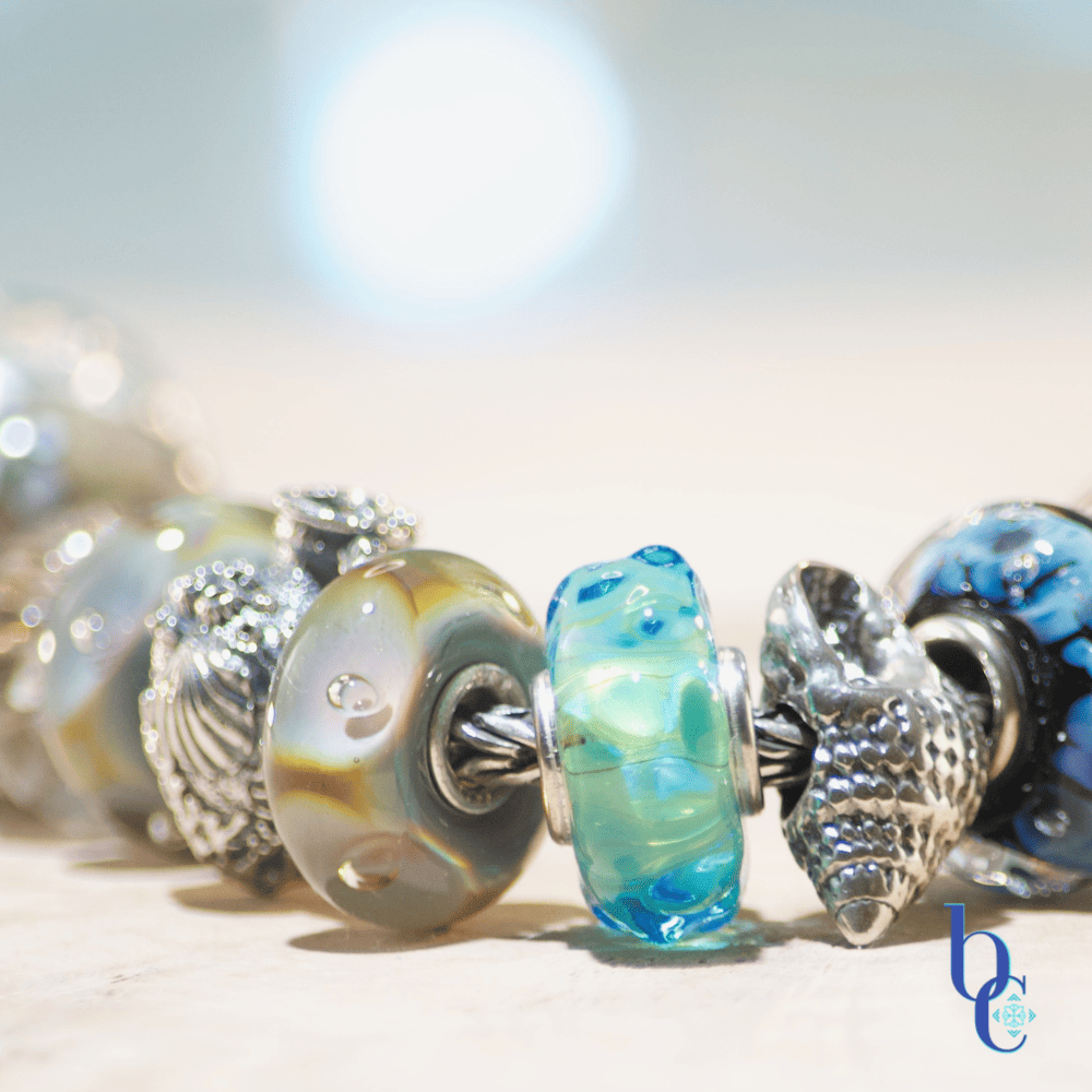 Close-up macro photograph of a European-style charm bracelet resting on a light surface. The focal beads include a textured silver nautilus shell, an aqua blue glass bead with internal bubbles, and the Kamane "Sandprint" bead featuring dark paw prints over a swirl of sandy beige and blue glass. The background is a soft, out-of-focus watercolor blue with bokeh lighting.