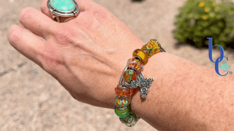 A woman's wrist wearing an ombré Trollbeads charm bracelet featuring retro floral glass beads and a silver butterfly dangle, photographed outdoors in natural lighting.