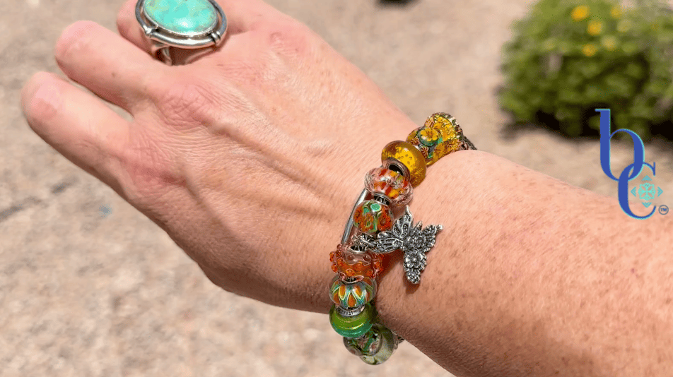 A woman's wrist wearing an ombré Trollbeads charm bracelet featuring retro floral glass beads and a silver butterfly dangle, photographed outdoors in natural lighting.