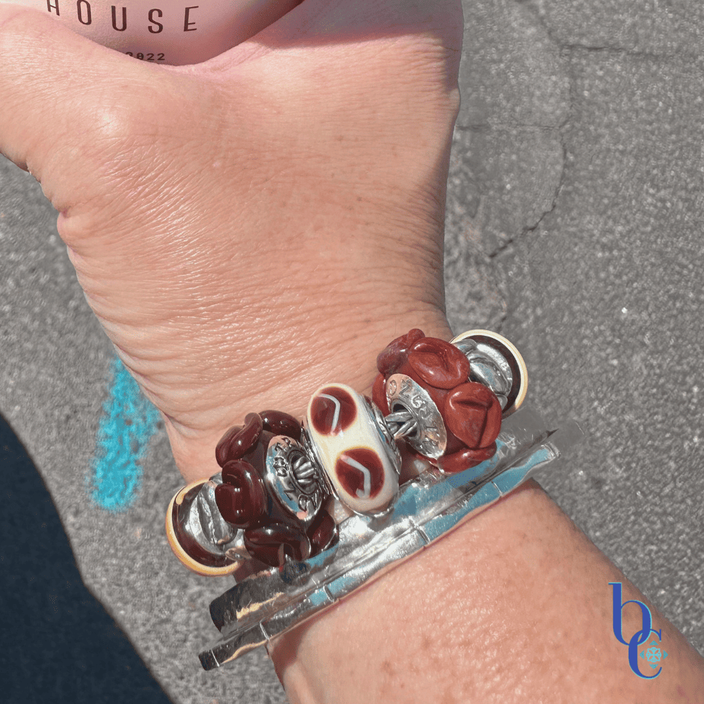 A woman's wrist wearing a chunky coffee-themed charm bracelet with TrueBeadz and Biagi Italy glass over a raw asphalt background.
