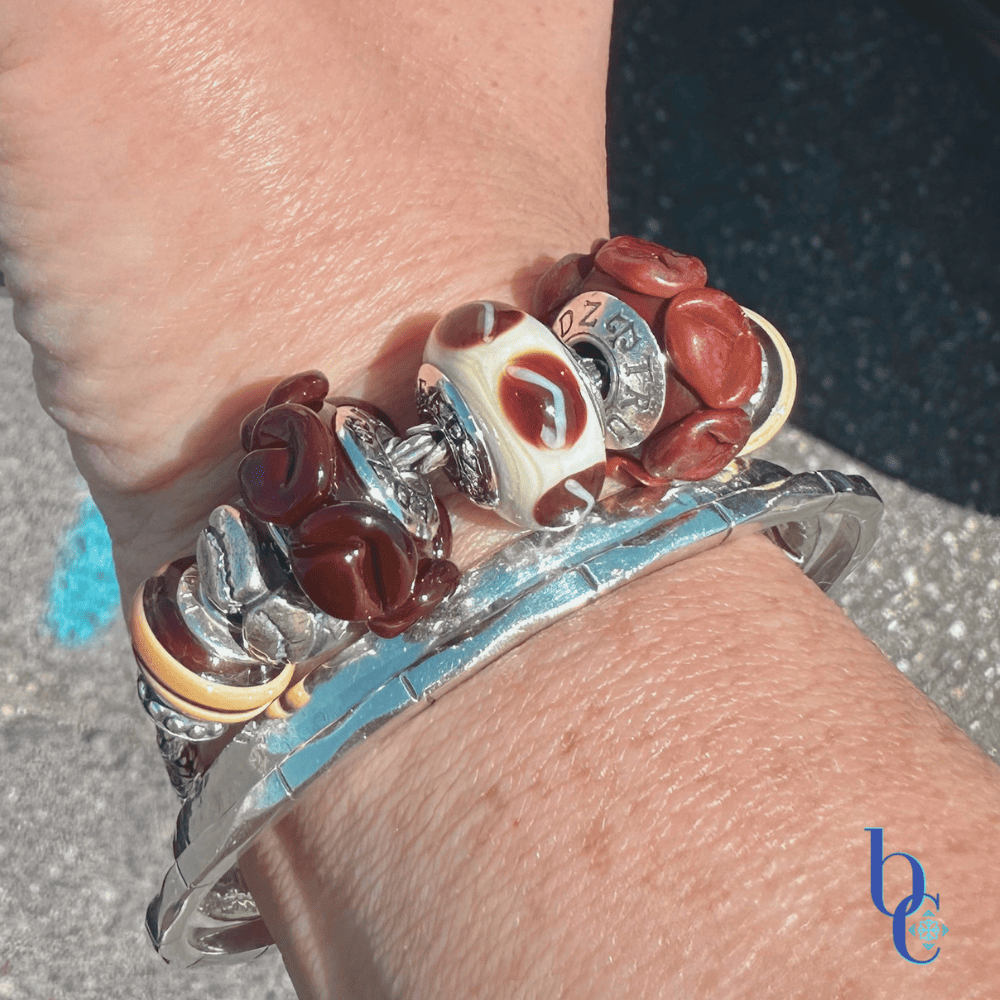 A woman's wrist wearing a chunky coffee-themed charm bracelet with TrueBeadz and Biagi Italy glass over a raw asphalt background.