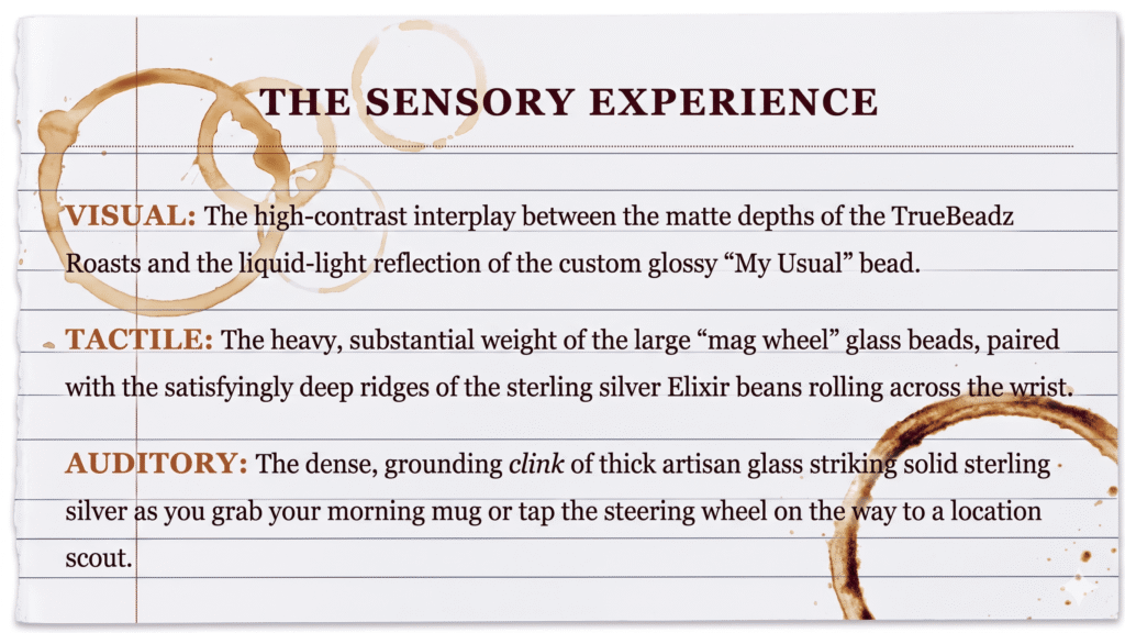 Text graphic designed to look like a sheet of lined notebook paper with brown coffee ring stains. The header reads "The Sensory Experience" in deep cabernet, followed by three sections: Visual, describing the high contrast between matte TrueBeadz Roasts and glossy beads; Tactile, detailing the heavy weight of glass "mag wheel" beads and textured sterling silver Elixir beans; and Auditory, describing the dense clink of artisan glass and solid silver against a morning coffee mug.