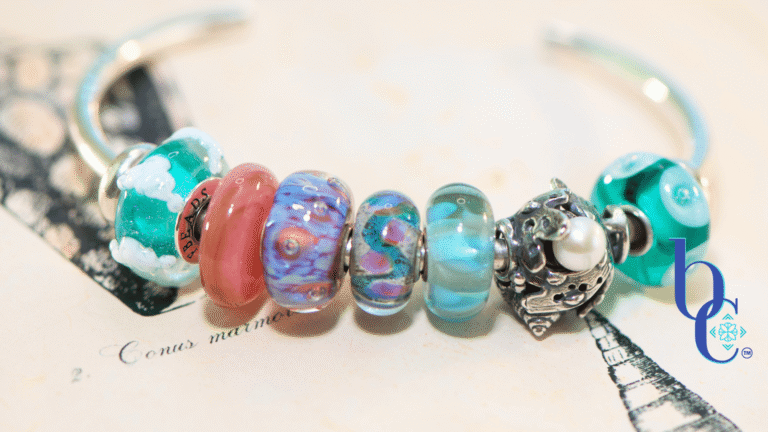 A macro photograph of a minimalist European charm bracelet on a polished silver bangle against a vintage book of shell engravings from Bermuda as a background. The arrangement features teal, aqua, and coral pink glass beads by Trollbeads and Elfbeads, centered around a detailed Trollbeads silver seashell and pearl charm.