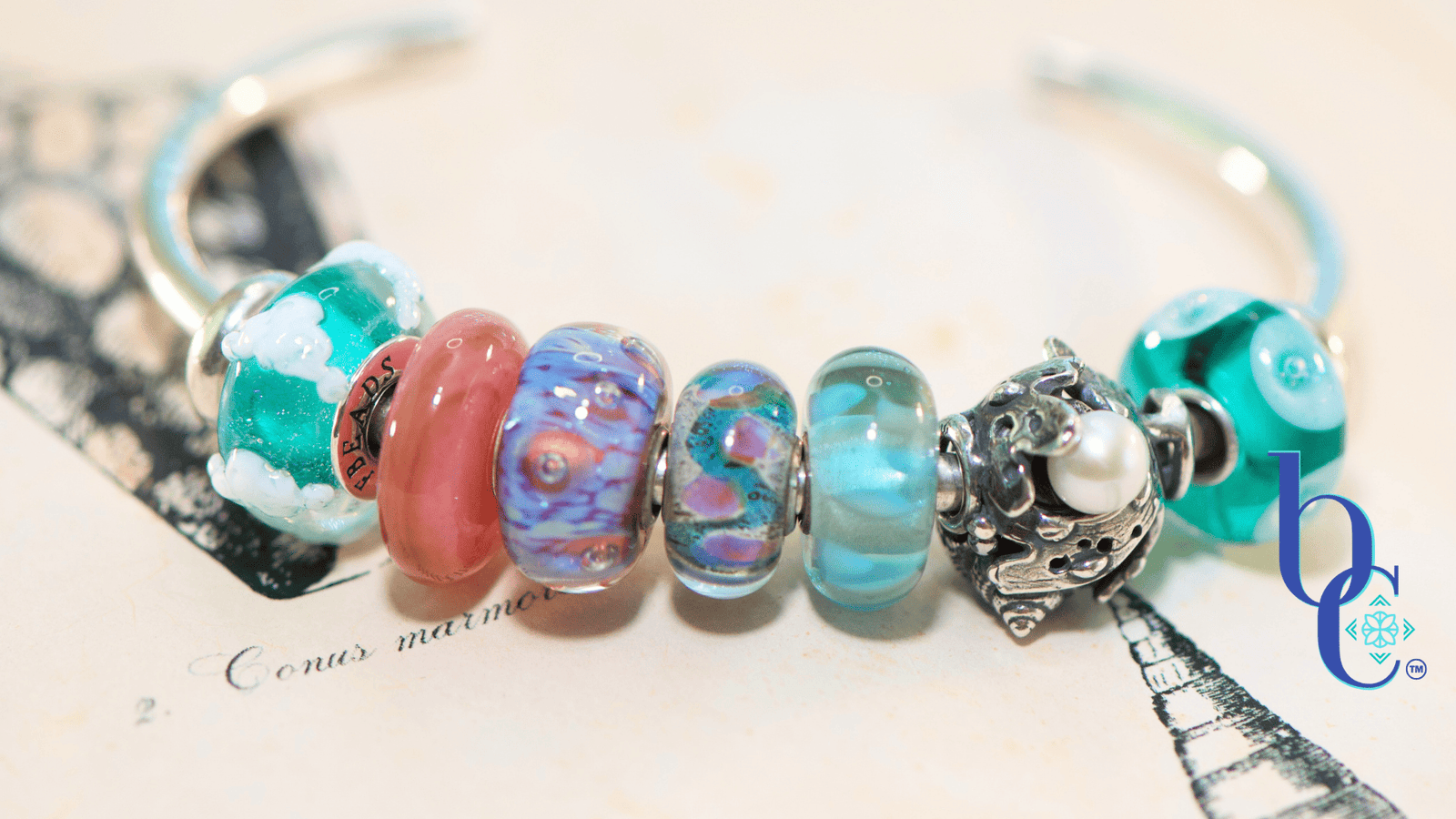 A macro photograph of a minimalist European charm bracelet on a polished silver bangle against a vintage book of shell engravings from Bermuda as a background. The arrangement features teal, aqua, and coral pink glass beads by Trollbeads and Elfbeads, centered around a detailed Trollbeads silver seashell and pearl charm.