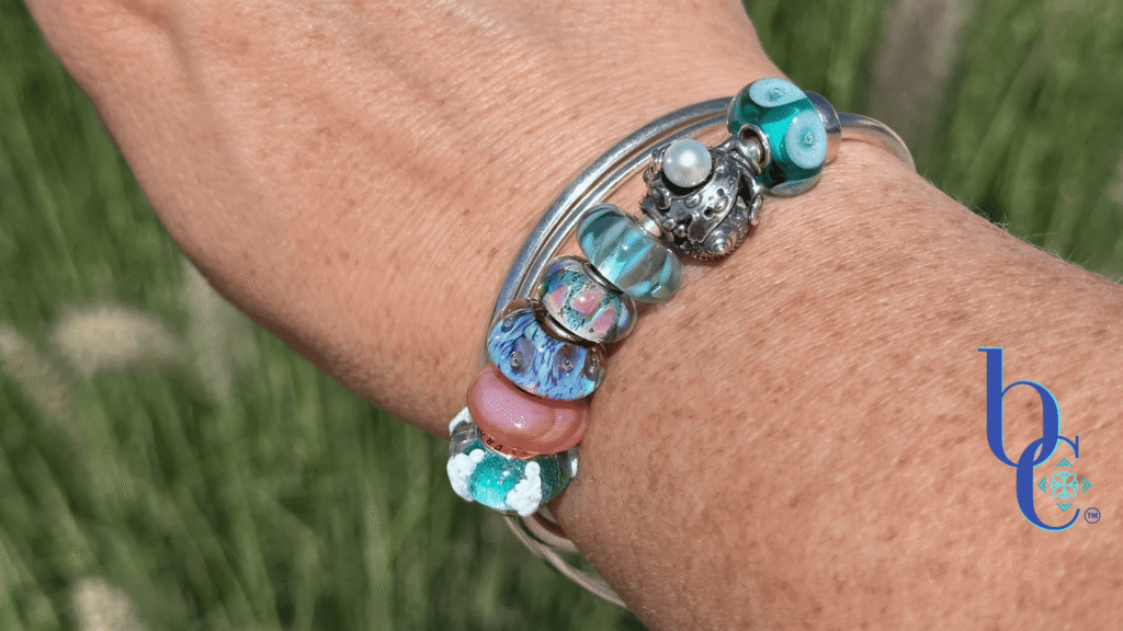 A macro photograph of a minimalist European charm bracelet on a polished silver bangle on a woman's wrist outside in natural lighting. The arrangement features teal, aqua, and coral pink glass beads by Trollbeads and Elfbeads, centered around a detailed Trollbeads silver seashell and pearl charm.