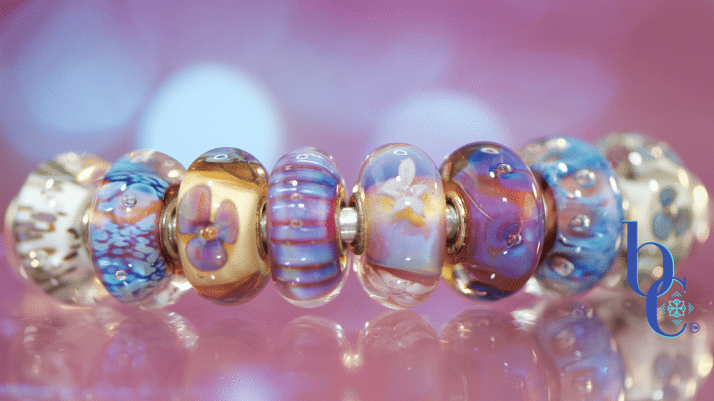 Close-up macro photograph of a glowing violet and blue European charm bracelet, featuring Trollbeads Sea Anemone and Purple Heaven Kit glass beads on a sterling silver chain, resting on an iridescent purple surface with bokeh lighting.
