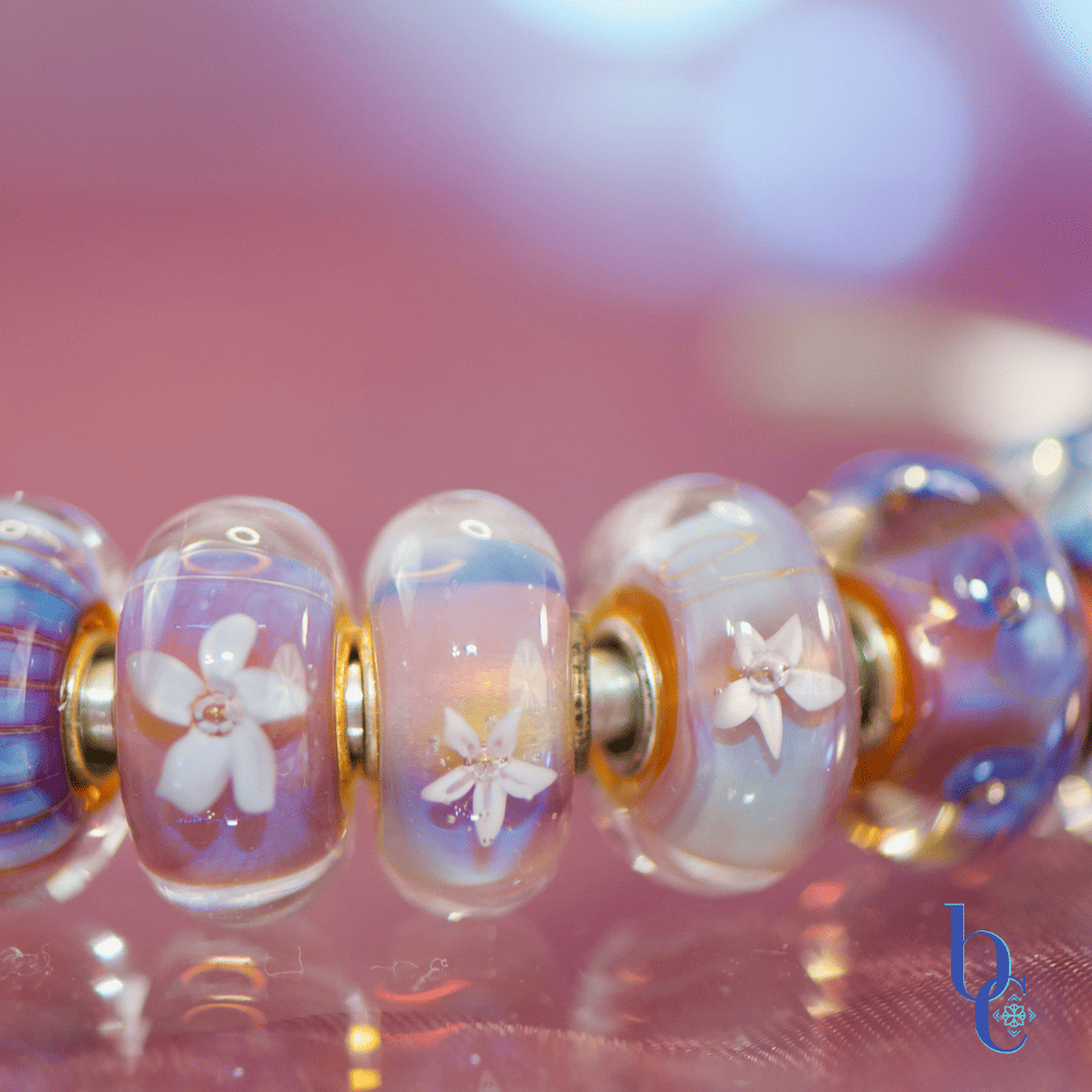 A macro studio shot of a vibrant purple and blue charm bracelet illuminated against a bokeh background, highlighting the details of retired artisan glass charms.
