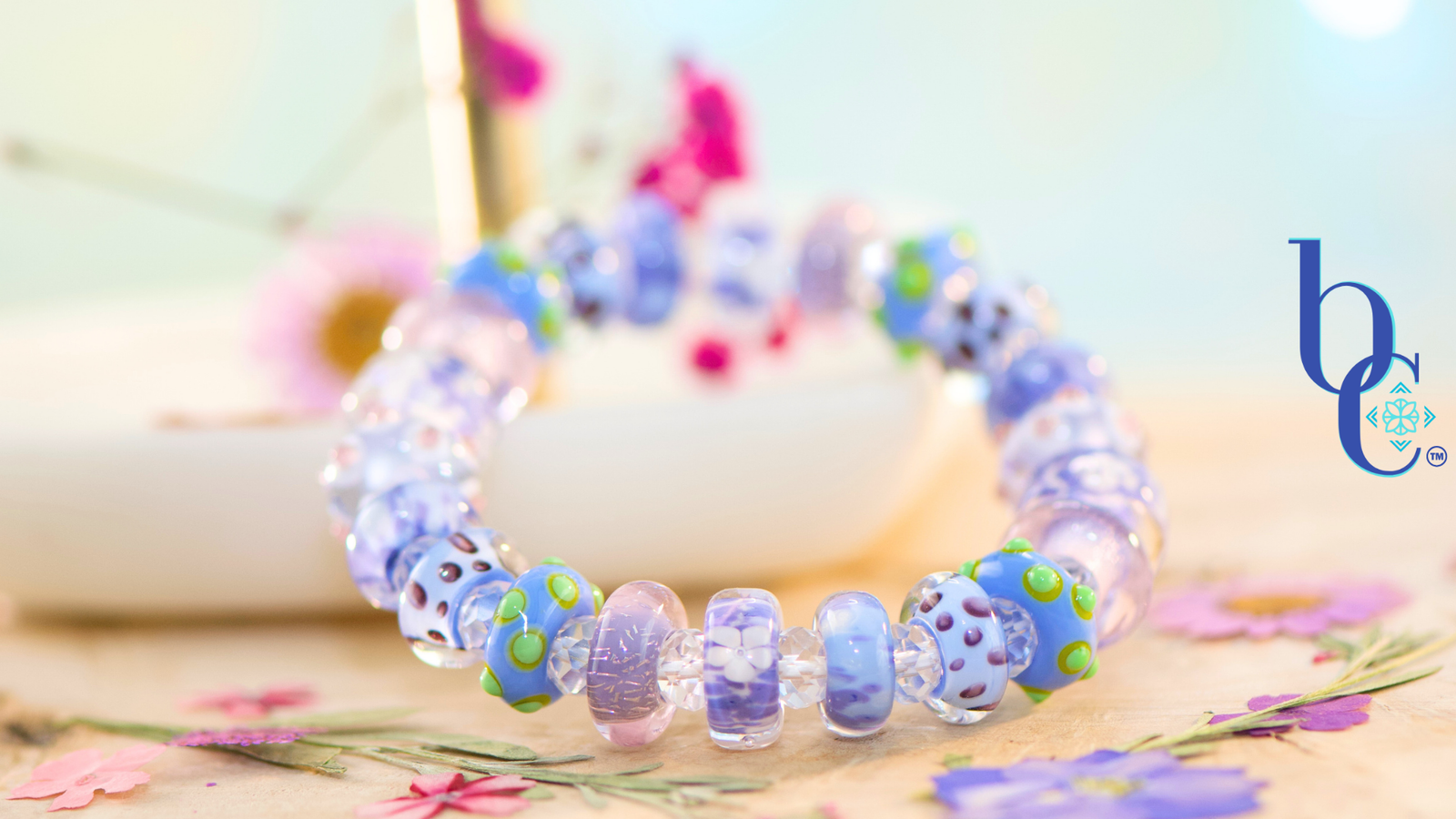 A macro shot of a handcrafted stretch bracelet resting on weathered driftwood, featuring uncored glass beads in vibrant shades of periwinkle, lavender, and violet with raised lime-green dots, spaced by clear faceted beads, nestled next to a bright pressed flowers.