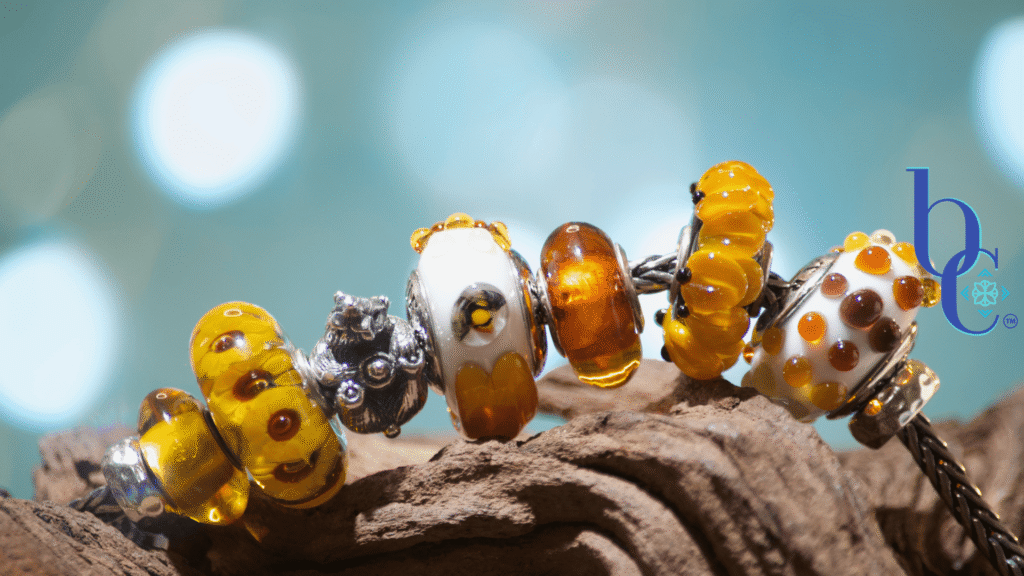 Extreme close-up of a European charm bracelet featuring a sculpted silver Black Raven honey bear charm nestled alongside translucent True Beadz honey glass beads, some natural amber beads, and a textured Kamane glass sunflower bead.