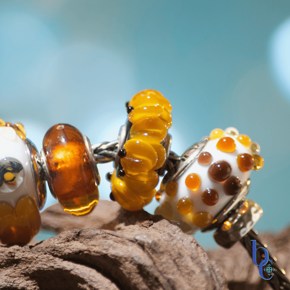 Extreme close-up of a European charm bracelet featuring a highly textured Kamane sunflower bead alongside a translucent Trollbeads cognac amber bead and a True Beadz honey colored dotty glass bead.