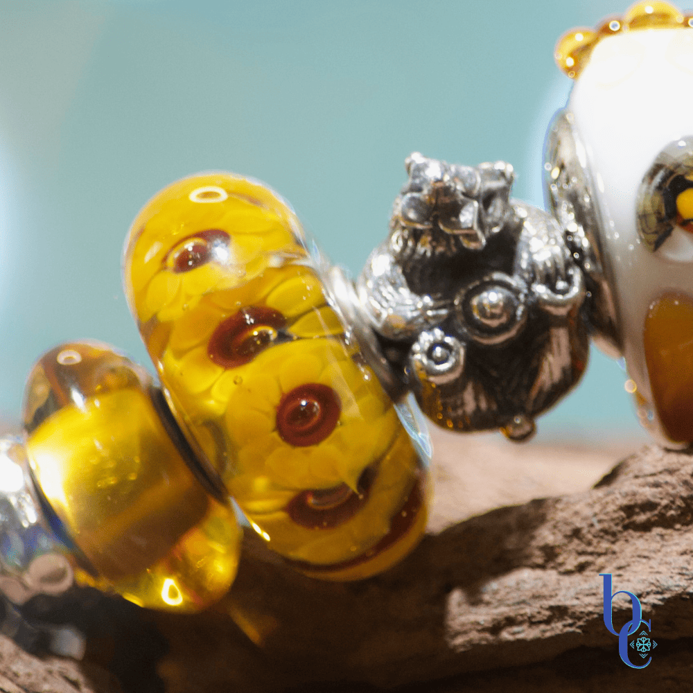 Extreme close-up of a European charm bracelet featuring a highly detailed jumbo Kamane sunflower bead alongside a honey colored Trollbeads amber bead.
