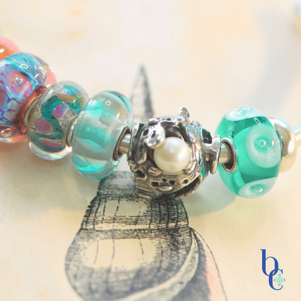 A macro photograph of a minimalist European charm bracelet on a polished silver bangle against a vintage book of shell engravings from Bermuda as a background. The arrangement features teal, aqua, and coral pink glass beads by Trollbeads and Elfbeads, centered around a detailed Trollbeads silver seashell and pearl charm.