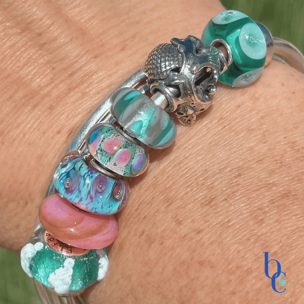 A macro photograph of a minimalist European charm bracelet on a polished silver bangle on a woman's wrist outside in natural lighting. The arrangement features teal, aqua, and coral pink glass beads by Trollbeads and Elfbeads, centered around a detailed Trollbeads silver seashell and pearl charm.