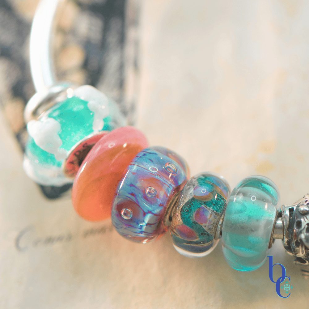 A macro photograph of a minimalist European charm bracelet on a polished silver bangle against a vintage book of shell engravings from Bermuda as a background. The arrangement features teal, aqua, and coral pink glass beads by Trollbeads and Elfbeads, centered around a detailed Trollbeads silver seashell and pearl charm.