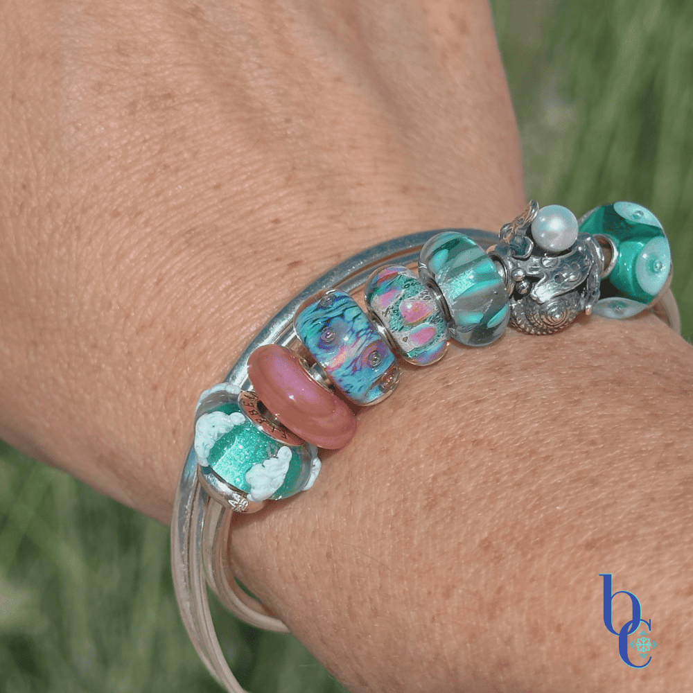A macro photograph of a minimalist European charm bracelet on a polished silver bangle on a woman's wrist outside in natural lighting. The arrangement features teal, aqua, and coral pink glass beads by Trollbeads and Elfbeads, centered around a detailed Trollbeads silver seashell and pearl charm.