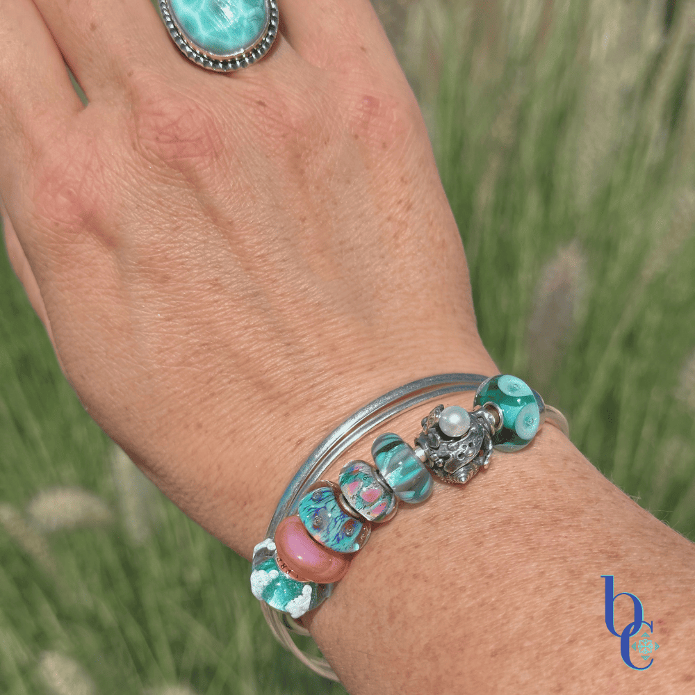 A macro photograph of a minimalist European charm bracelet on a polished silver bangle on a woman's wrist outside in natural lighting. The arrangement features teal, aqua, and coral pink glass beads by Trollbeads and Elfbeads, centered around a detailed Trollbeads silver seashell and pearl charm.