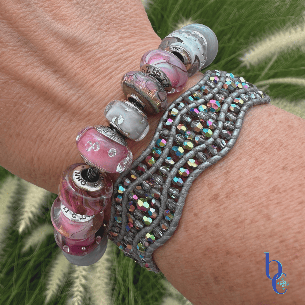 A close-up lifestyle shot of a gray and pink artisan glass charm bracelet catching the warm, natural sunlight on a wrist, showcasing the iridescent shimmer of the charms.
