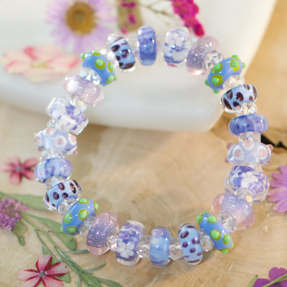 A macro flatlay shot of a handcrafted stretch bracelet resting on weathered driftwood, featuring uncored glass beads in vibrant shades of periwinkle, lavender, and violet with raised lime-green dots, spaced by clear faceted beads, nestled next to a bright pressed flowers.
