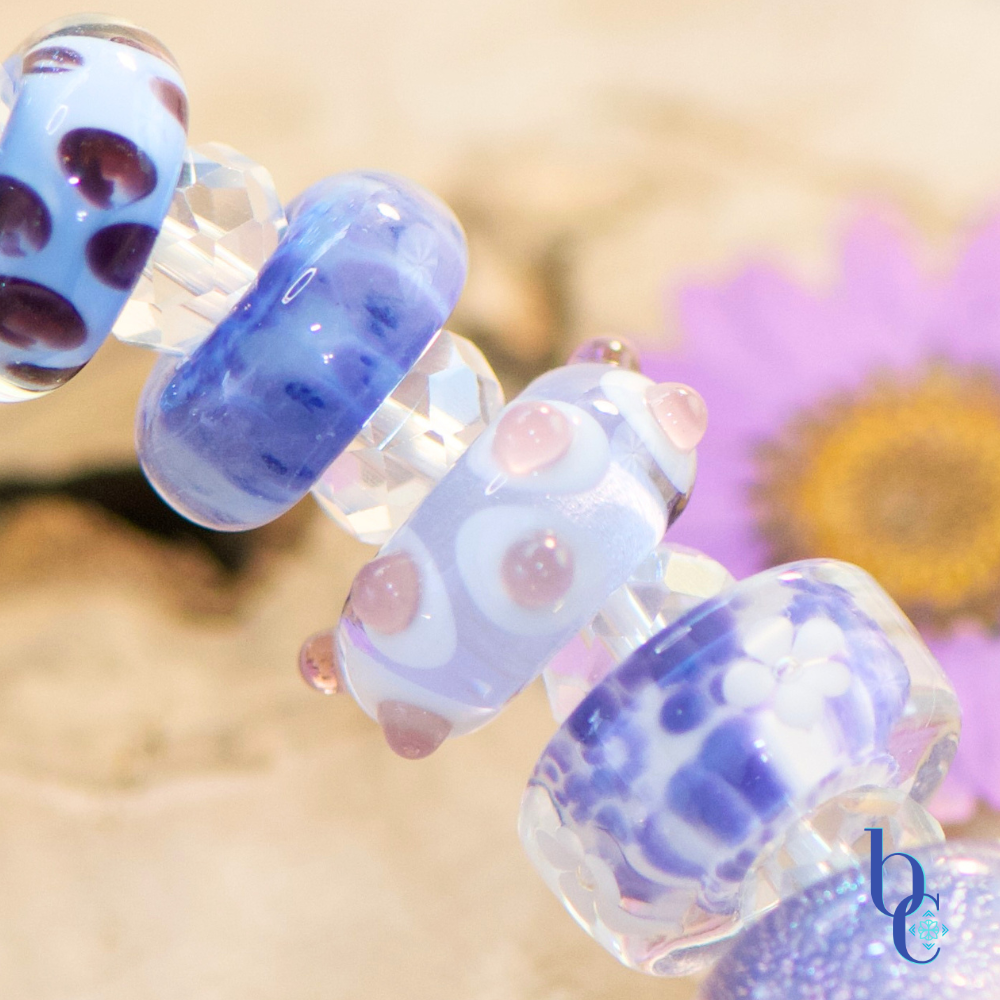 A macro shot of  handcrafted glass beads resting on weathered driftwood, featuring uncored glass beads in vibrant shades of periwinkle, lavender, and violet spaced by clear faceted beads, nestled next to a bright pressed flowers.