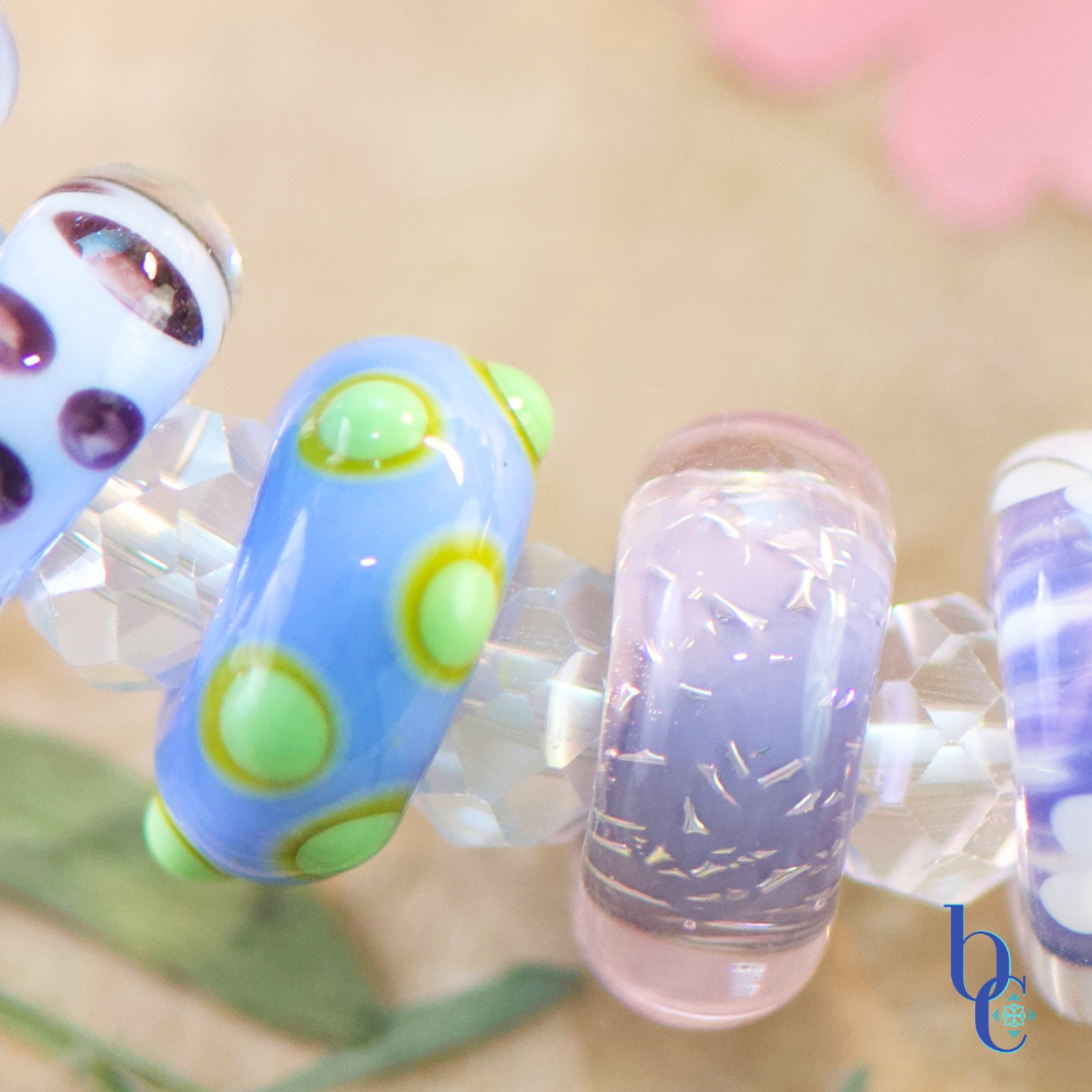 A macro shot of a handcrafted glass beads resting on weathered driftwood, featuring uncored glass beads in vibrant shades of periwinkle, lavender, and violet with raised lime-green dots, spaced by clear faceted beads, nestled next to a bright pressed flowers.