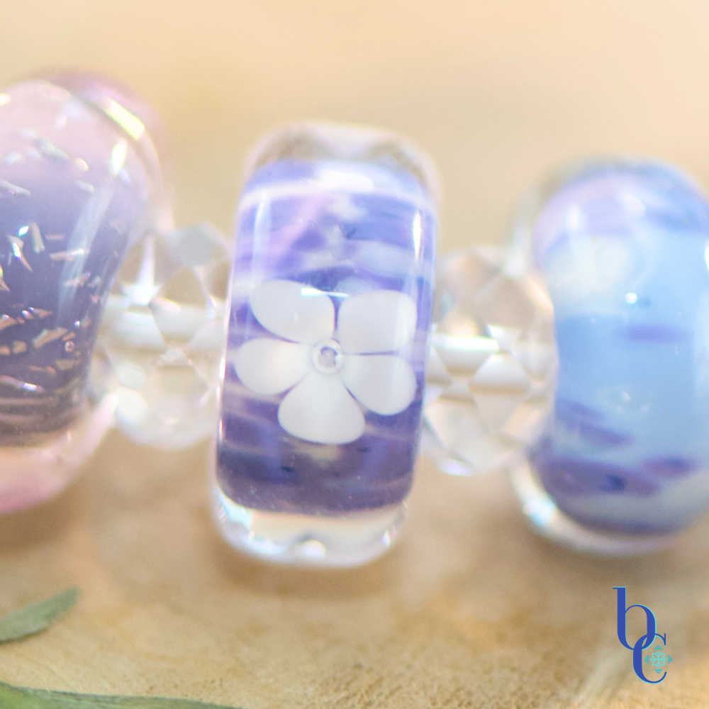 A macro shot of handcrafted glass beads resting on weathered driftwood, featuring uncored glass beads in vibrant shades of periwinkle, lavender, and violet spaced by clear faceted beads, nestled next to a bright pressed flowers.