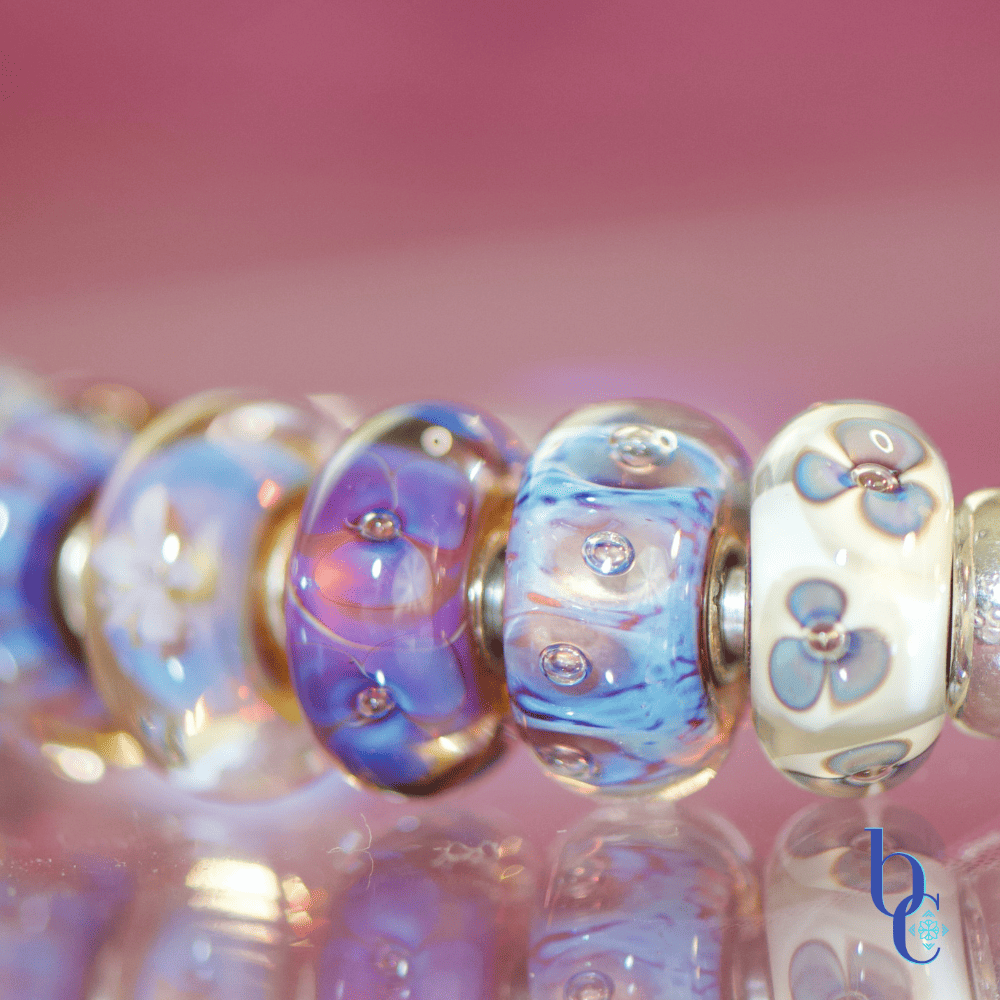 A macro studio shot of a vibrant purple and blue charm bracelet illuminated against a bokeh background, highlighting the details of retired artisan glass charms.
