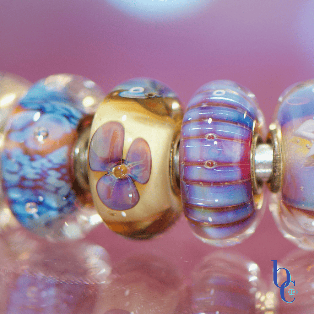 A macro studio shot of a vibrant purple and blue charm bracelet illuminated against a bokeh background, highlighting the details of retired artisan glass charms.
