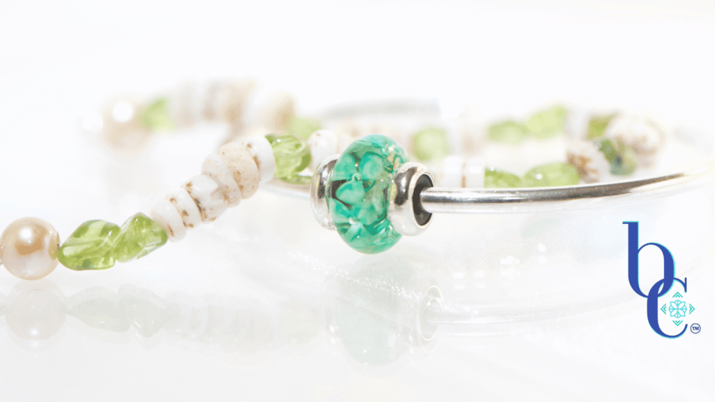 A minimalist luxury jewelry layout featuring a genuine white Hawaiian puka shell bracelet with green peridot nuggets next to a polished silver Trollbeads bangle holding a single Forest Anemone glass bead on a glowing white surface.