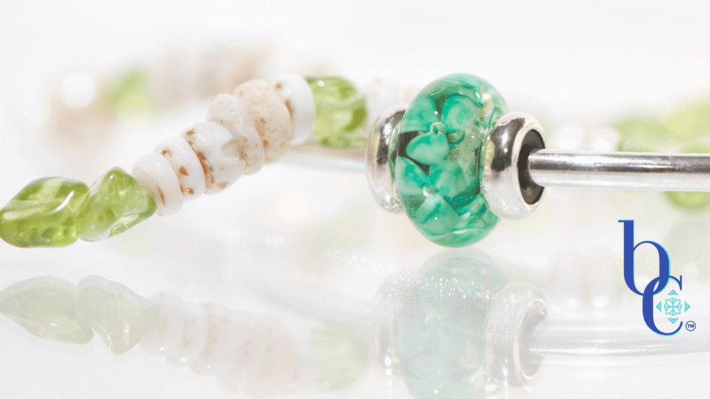 A minimalist luxury jewelry layout featuring a genuine white Hawaiian puka shell bracelet with green peridot nuggets next to a polished silver Trollbeads bangle holding a single Forest Anemone glass bead on a glowing white surface.