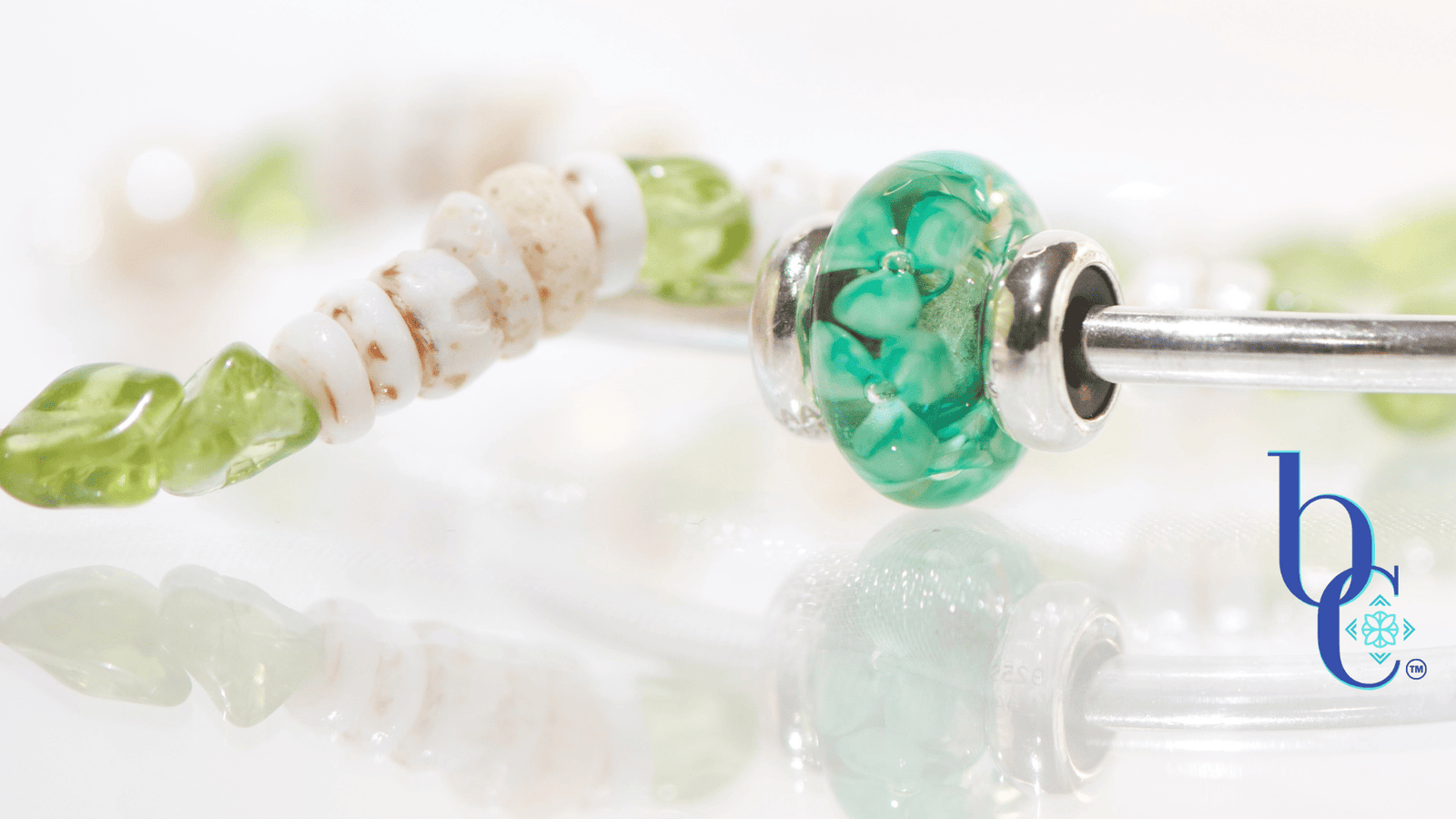 A minimalist luxury jewelry layout featuring a genuine white Hawaiian puka shell bracelet with green peridot nuggets next to a polished silver Trollbeads bangle holding a single Forest Anemone glass bead on a glowing white surface.