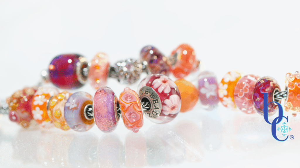 High-contrast macro photograph of a European-style charm bracelet featuring bright orange, hot pink, and purple artisan glass beads interspersed with highly textured silver floral charms, resting on an underlit plexi base.