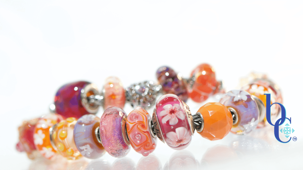 High-contrast macro photograph of a European-style charm bracelet featuring bright orange, hot pink, and purple artisan glass beads interspersed with highly textured silver floral charms, resting on an underlit plexi base.