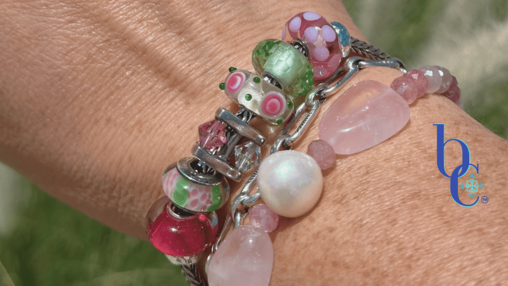 High-contrast macro photograph of a European-style charm bracelet featuring pink and green artisan glass beads interspersed with a rotating crystal charm, resting on woman's wrist in outdoor light.