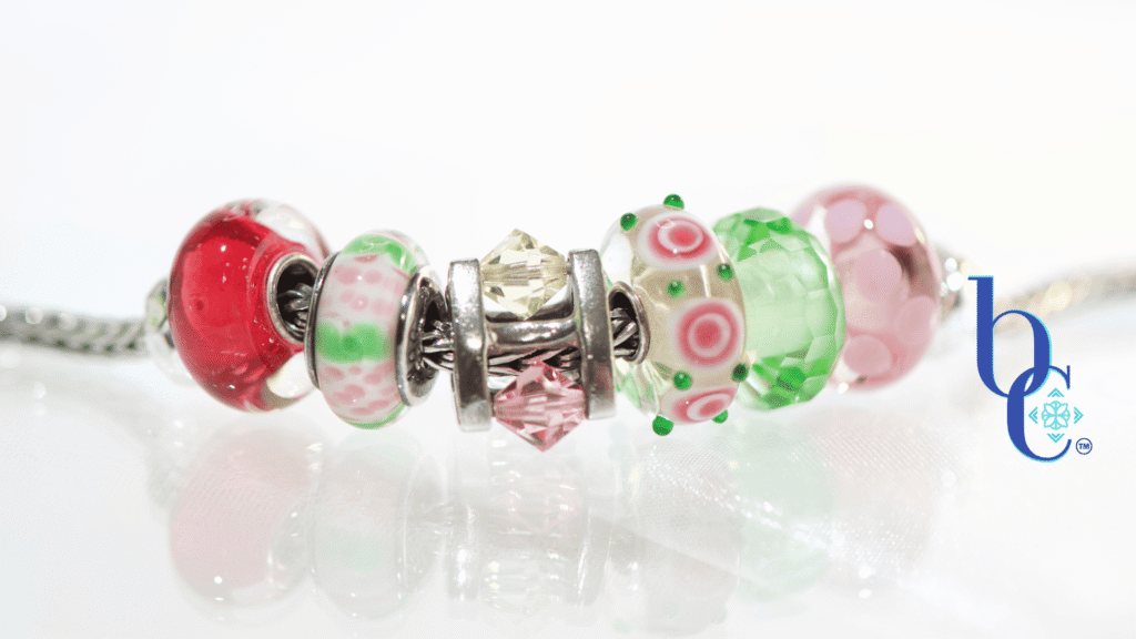 High-contrast macro photograph of a European-style charm bracelet featuring pink and green artisan glass beads interspersed with a rotating crystal charm, resting on an underlit plexi base.