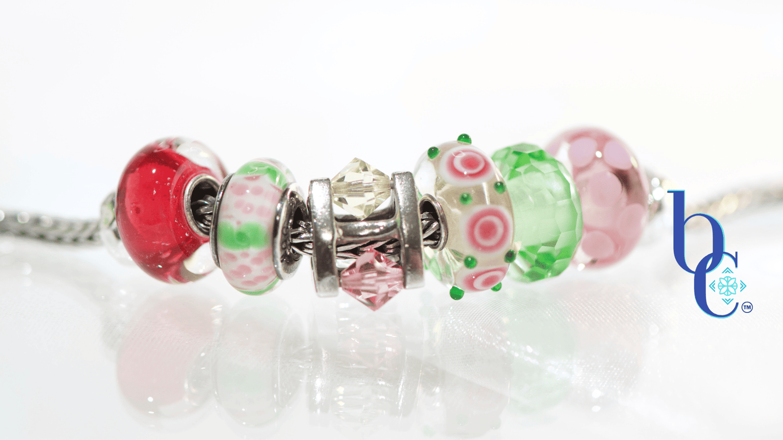 High-contrast macro photograph of a European-style charm bracelet featuring pink and green artisan glass beads interspersed with a rotating crystal charm, resting on an underlit plexi base.