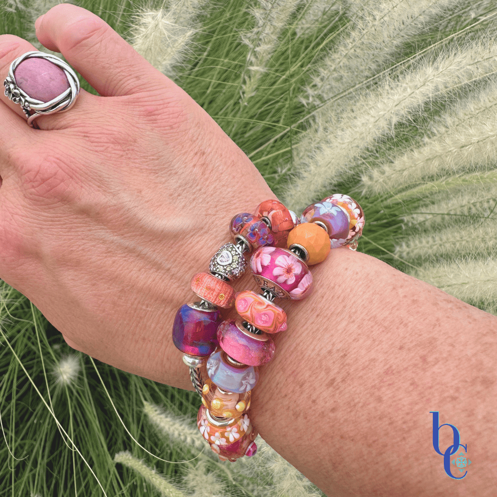 A woman's wrist wearing a heavily stacked charm bracelet with vibrant tangerine and magenta glass beads, catching the bright, natural outdoor sunlight against a blurred summer background.