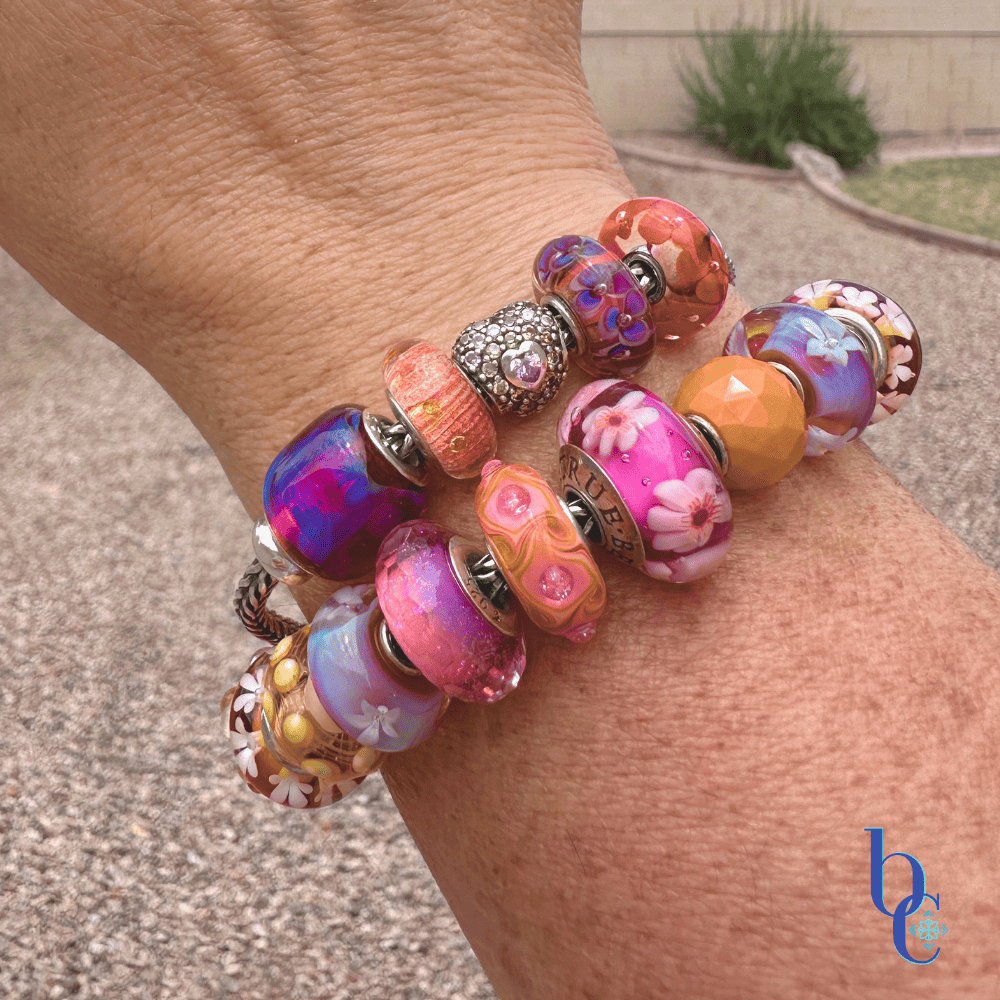 A woman's wrist wearing a heavily stacked charm bracelet with vibrant tangerine and magenta glass beads, catching the bright, natural outdoor sunlight against a blurred summer background.