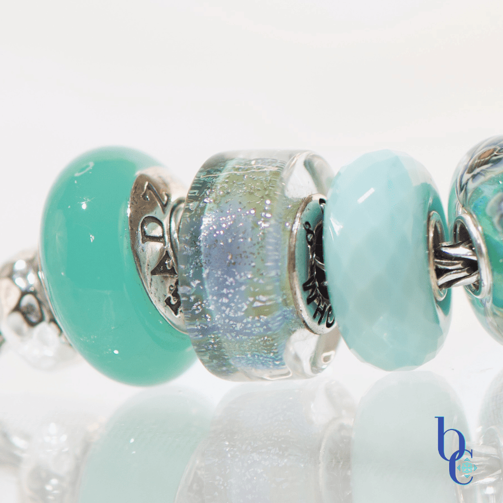 A macro photograph of a European charm bracelet resting on a highly reflective white surface. The stack features nine glass beads in saturated tones of sage green, icy mint, and vibrant teal, including a focal Elfbeads Azure glass bead. The background is a soft, luminous white with bokeh light circles.