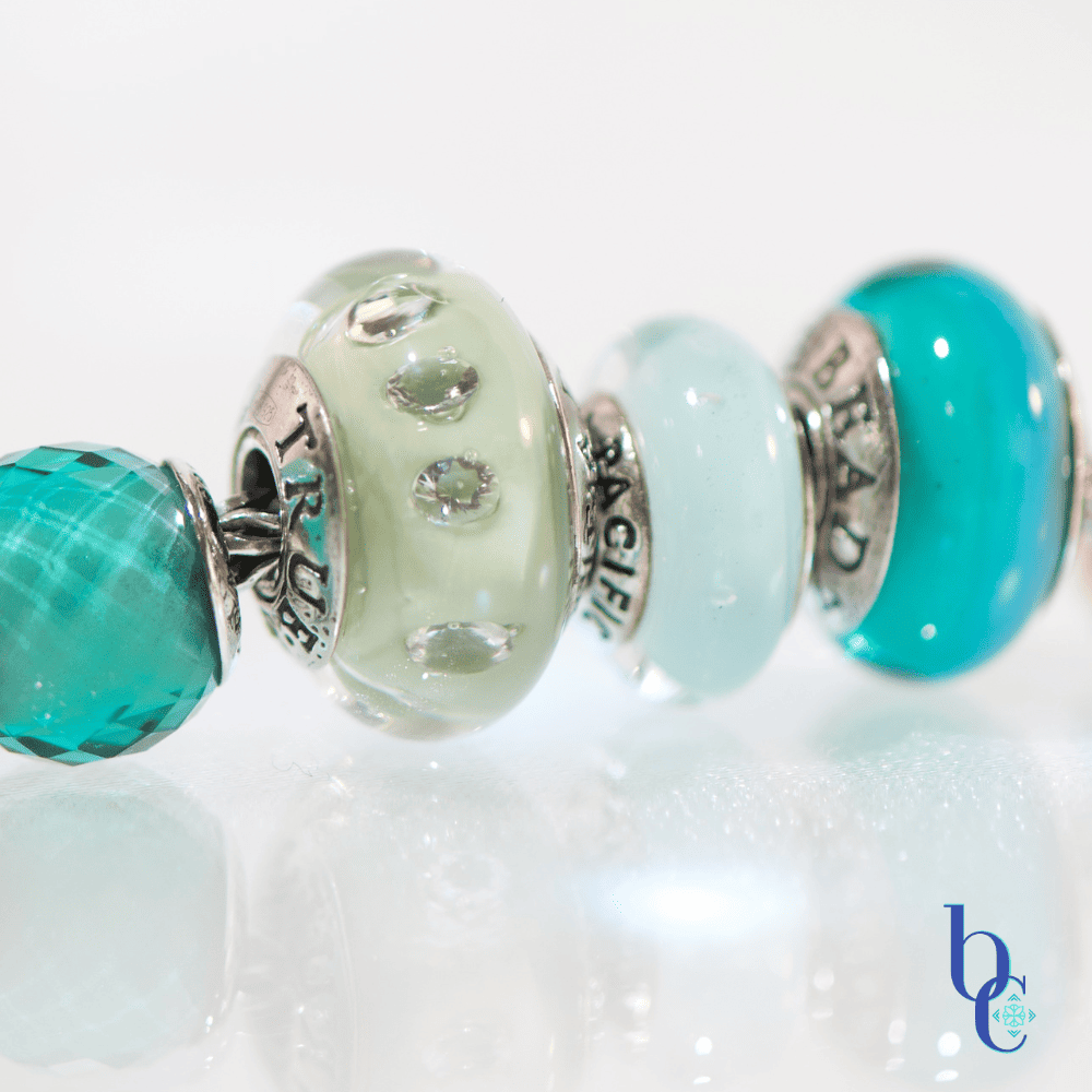 A macro photograph of a European charm bracelet resting on a highly reflective white surface. The stack features nine glass beads in saturated tones of sage green, icy mint, and vibrant teal, including a focal Elfbeads Azure glass bead. The background is a soft, luminous white with bokeh light circles.
