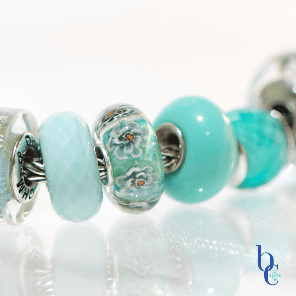 A macro photograph of a European charm bracelet resting on a highly reflective white surface. The stack features nine glass beads in saturated tones of sage green, icy mint, and vibrant teal, including a focal Elfbeads Azure glass bead. The background is a soft, luminous white with bokeh light circles.