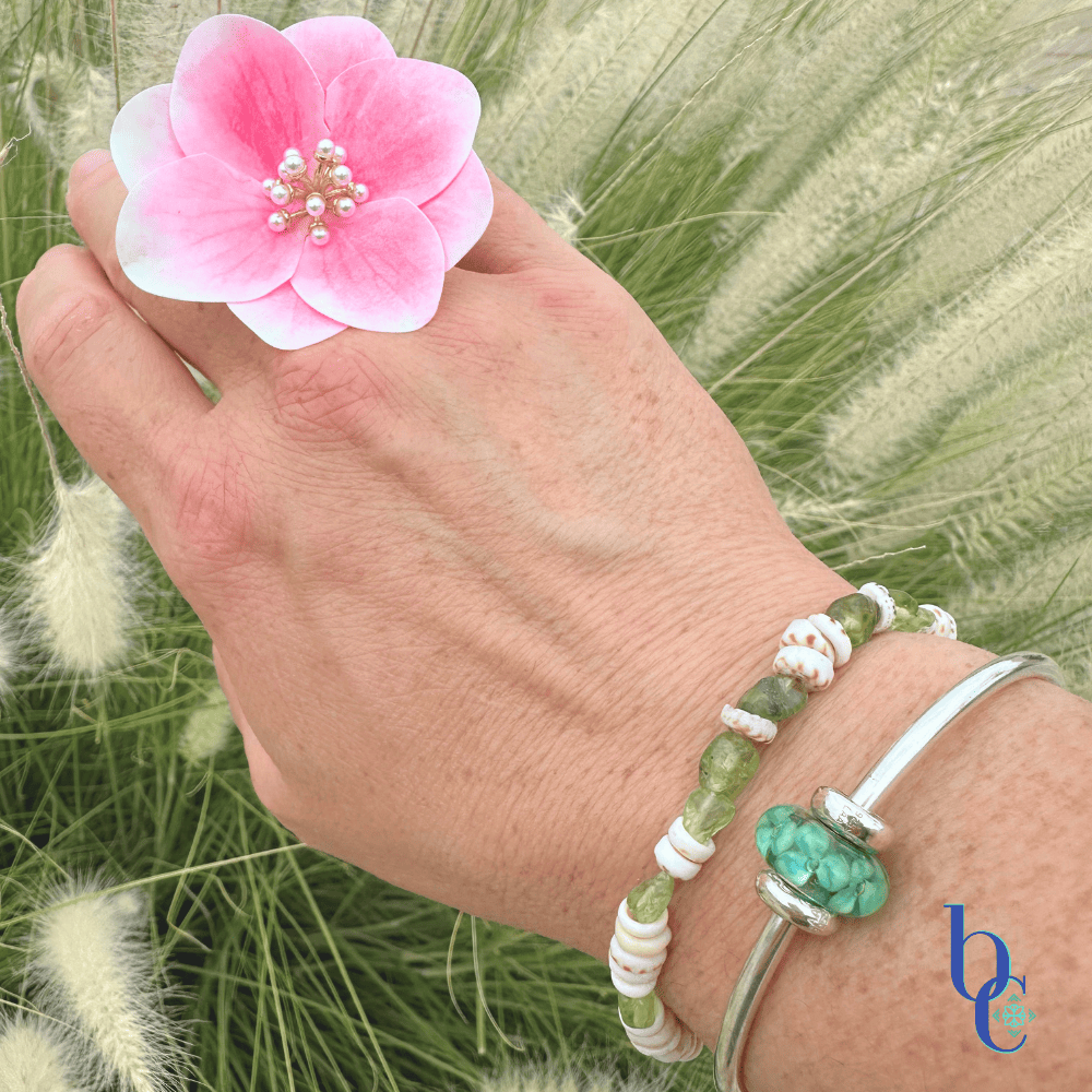 A wrist stack showing a natural Hawaiian puka shell and peridot gemstone bracelet layered with a sleek sterling silver Trollbeads bangle featuring the Forest Anemone bead under bright, warm sunlight on a woman’s wrist.