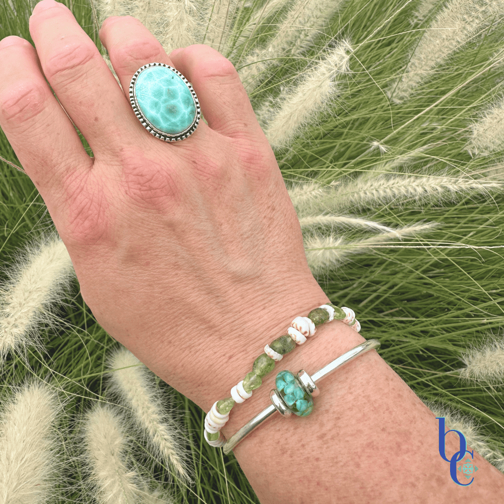 A wrist stack showing a natural Hawaiian puka shell and peridot gemstone bracelet layered with a sleek sterling silver Trollbeads bangle featuring the Forest Anemone bead under bright, warm sunlight on a woman’s wrist.