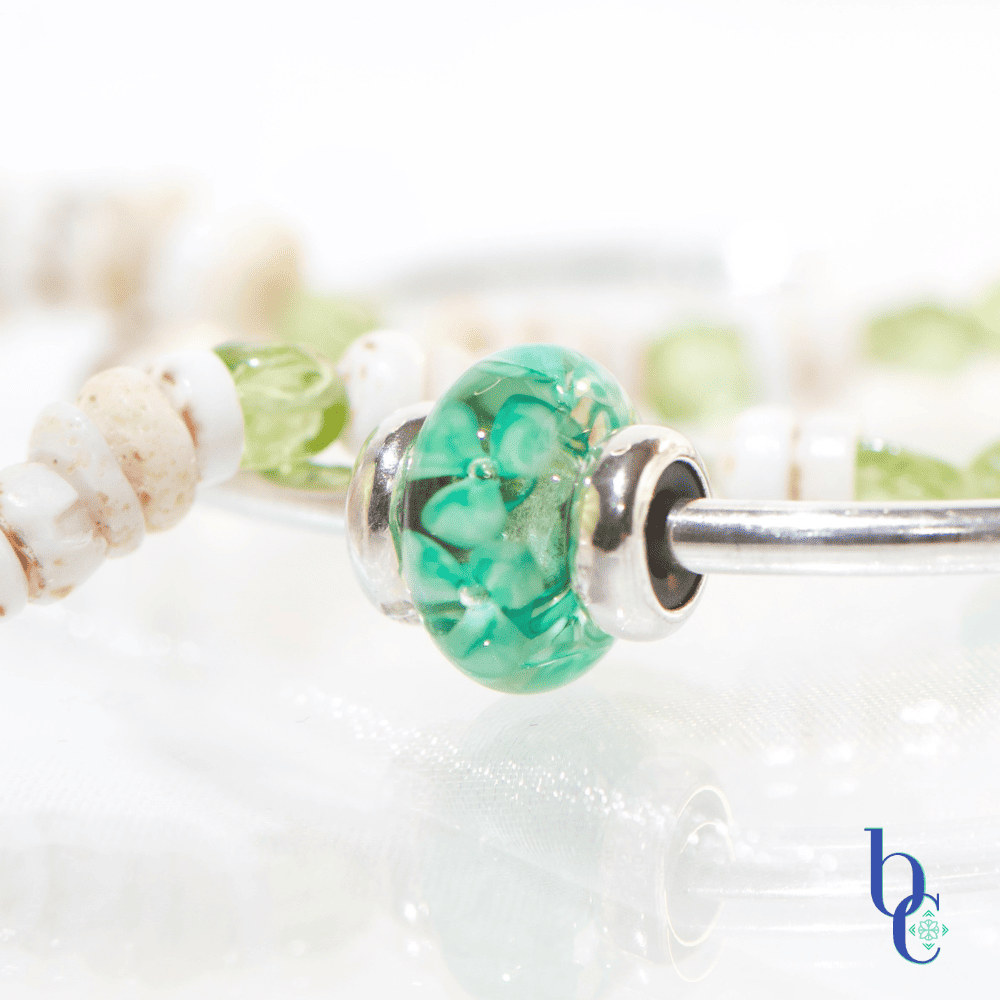 A macro of the Trollbeads Forest Anemone in a minimalist luxury jewelry layout featuring a genuine white Hawaiian puka shell bracelet with green peridot nuggets next to a polished silver Trollbeads bangle holding a single Forest Anemone glass bead on a glowing white surface.