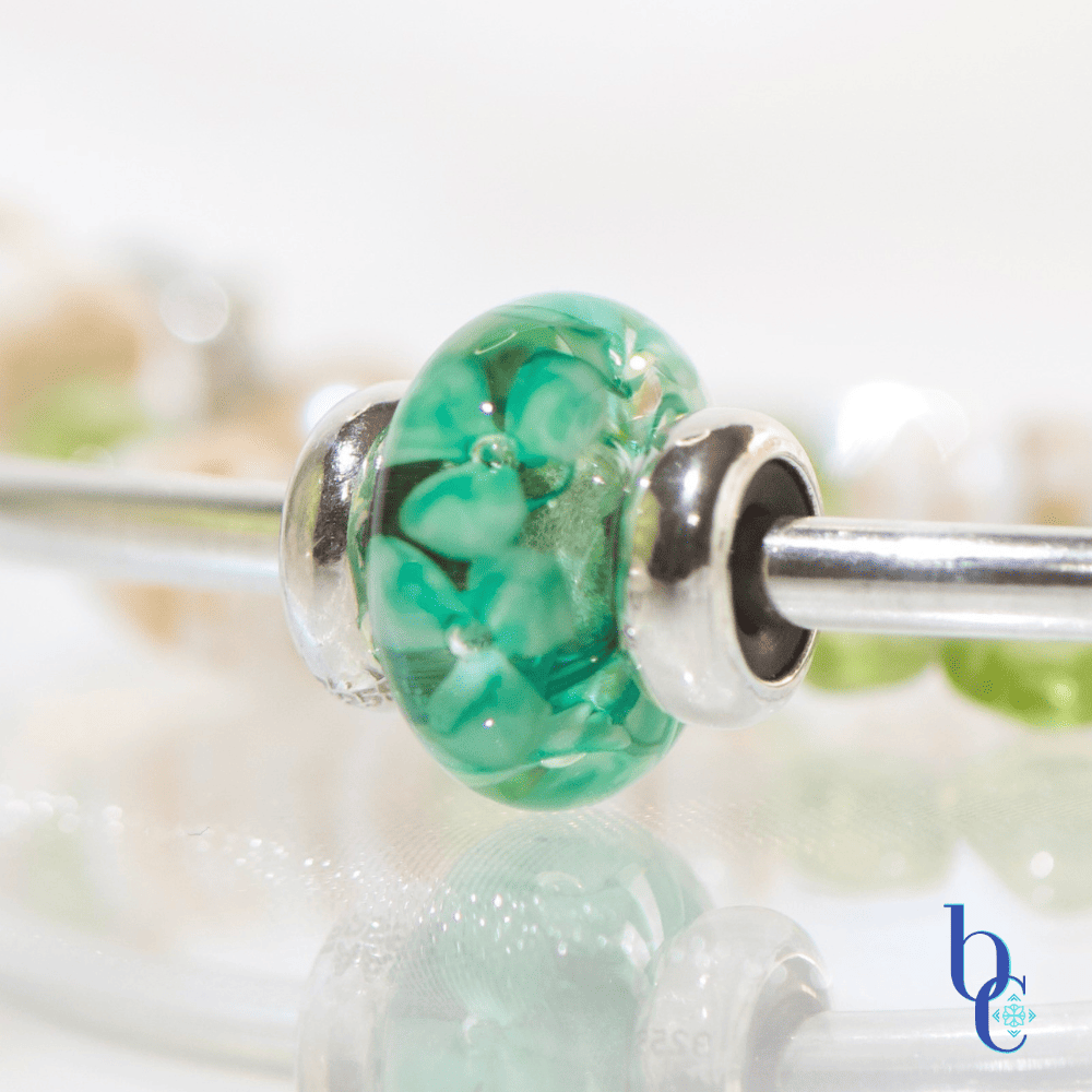 A macro of the Trollbeads Forest Anemone in a minimalist luxury jewelry layout featuring a genuine white Hawaiian puka shell bracelet with green peridot nuggets next to a polished silver Trollbeads bangle holding a single Forest Anemone glass bead on a glowing white surface.
