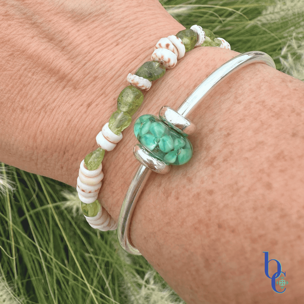 A wrist stack showing a natural Hawaiian puka shell and peridot gemstone bracelet layered with a sleek sterling silver Trollbeads bangle featuring the Forest Anemone bead under bright, warm sunlight on a woman’s wrist.